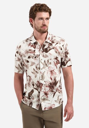 Man met kort bruin haar en baard, gekleed in een beige en bruine bloemenprint kort mouw overhemd met knopen en een bruine broek, staand met één hand in de zak.