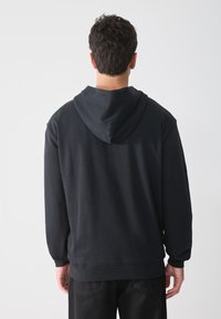 Sudadera con capucha negra hecha de una tela suave, con un corte relajado, mangas largas y un dobladillo acanalado. Muestra la vista trasera y una textura lisa.