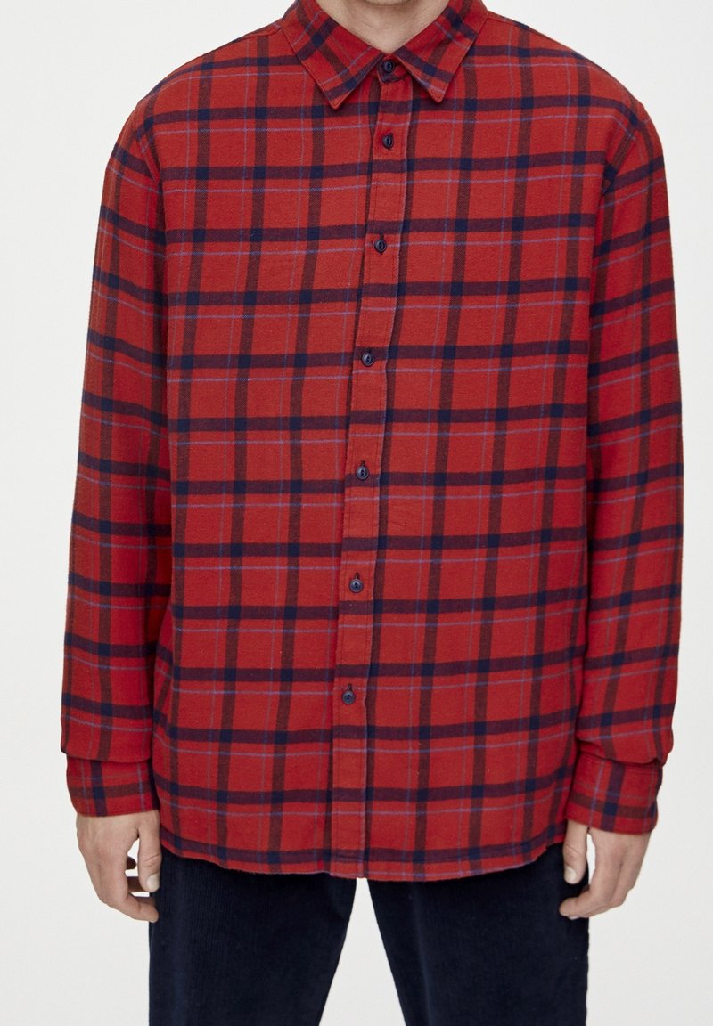 Homme portant une chemise à carreaux rouge et noir à manches longues avec col, debout devant un fond uni.