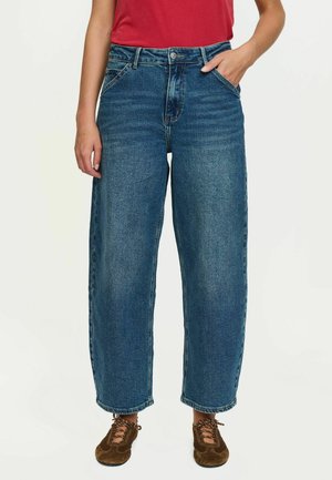 Personne portant un jean bleu taille haute à jambes larges et des chaussures marron à lacets, avec une main dans la poche et un haut rouge visible.