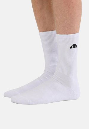 Paar weiße Crew-Socken mit schwarzem Logo am Knöchel, getragen an bloßen Beinen, vor weißem Hintergrund gezeigt.