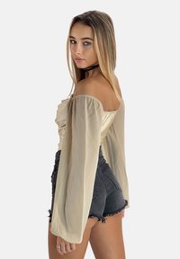 Beige schulterfreies Oberteil mit langen transparenten Ärmeln, kombiniert mit hoch geschnittenen schwarzen Denim-Shorts mit ausgefransten Kanten.