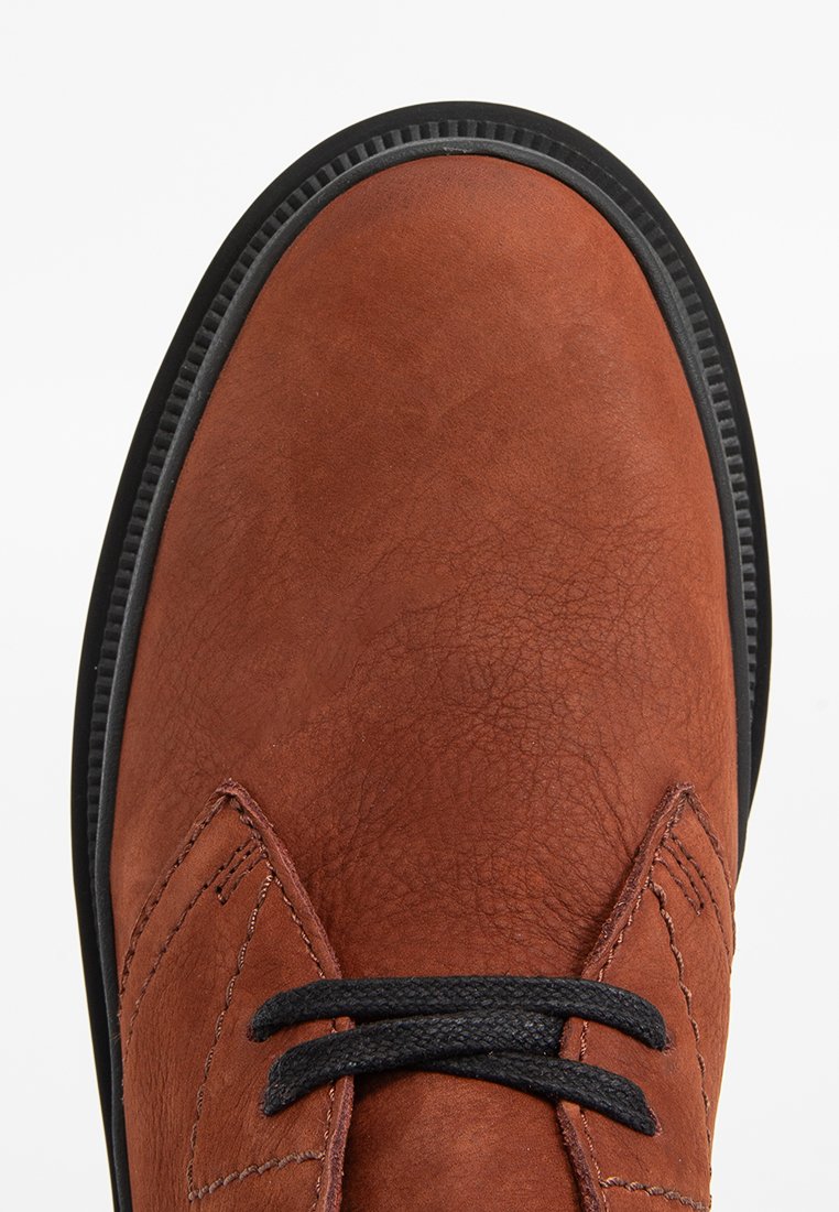 Chaussure en cuir marron avec une finition texturée, présentant une semelle en caoutchouc noir et des lacets, ainsi que des détails cousus sur les côtés.