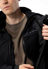 Černá zipová bunda s fleecovou podšívkou, obsahující síťovanou vnitřní kapsu. Nositá přes světle hnědý tričko, s viditelným logem na vnitřním límci.