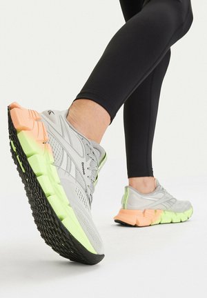 Osoba w szarych i neonowych butach do biegania Reebok oraz czarnych legginsach, unosząca jedną stopę, aby pokazać teksturowaną podeszwę i wzór buta.