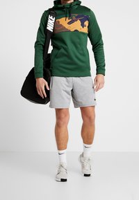 Homme portant un sweat à capuche vert avec un panneau abstrait, un short Nike gris, des chaussettes blanches Nike et des baskets, portant un sac de sport Nike noir à l'épaule.