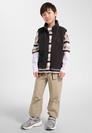 Jonge jongen staat, draagt een gestreept shirt met lange mouwen, een zwart fleecevest, beige cargobroek en grijze sneakers tegen een effen witte achtergrond.