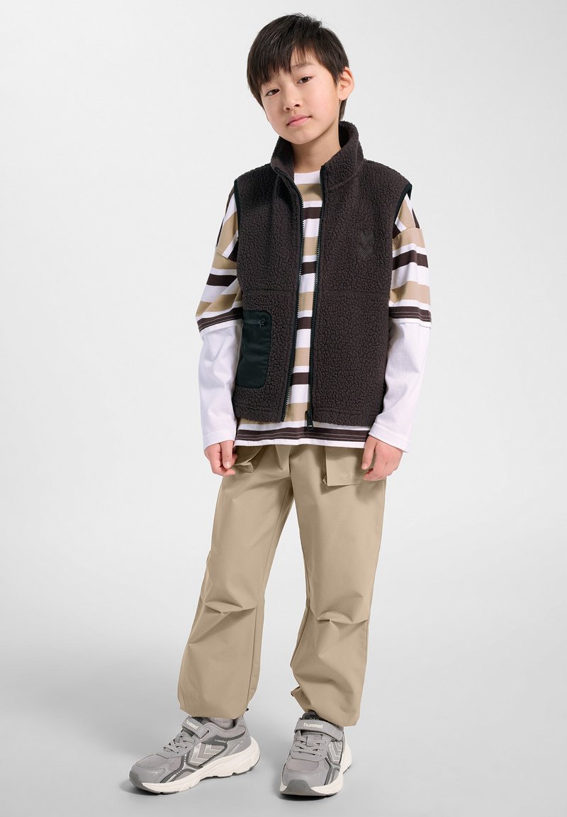 Młody chłopiec stoi, ubrany w pasiastą koszulę z długim rękawem, czarną kamizelkę polarową, beżowe spodnie typu cargo i szare trampki na jednolitym białym tle.
