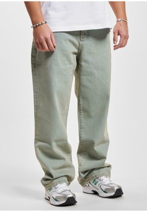 Person trägt lockere hellgrüne Jeans, weiße Sneakers mit grünen Akzenten und ein weißes T-Shirt, steht mit entspannten Händen entlang der Seiten.