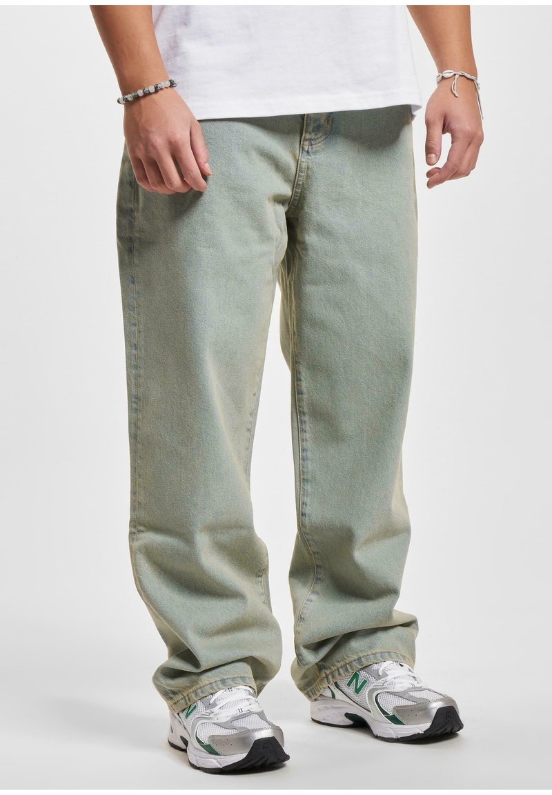 Personne portant un jean ample vert clair, des baskets blanches avec des touches vertes et un t-shirt blanc, debout les mains détendues le long du corps.