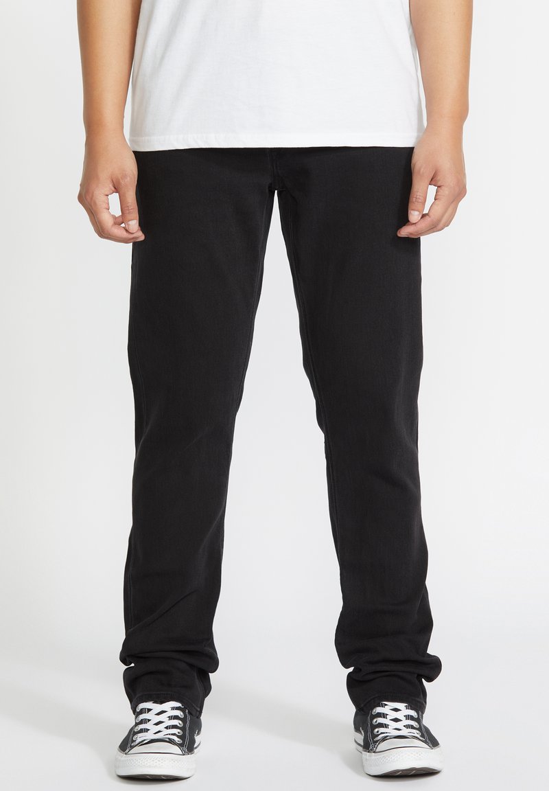 Personne portant un pantalon noir, une chemise blanche et des baskets décontractées noires et blanches, debout avec les mains détendues le long du corps.