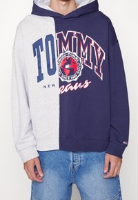 Sweatshirt met capuchon, linkerzijde grijs en rechterzijde marineblauw, met "TOMMY" tekst en wereldbolafbeelding in rood en wit. Gemaakt van katoenmix.
