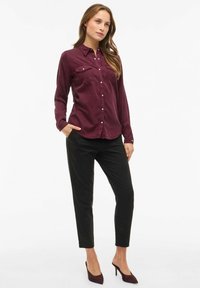 Burgundowa koszula z długim rękawem i dwoma kieszeniami na piersi, zapinana na guziki, w połączeniu z czarnymi spodniami typu slim-fit i ciemnofioletowymi szpilkami.