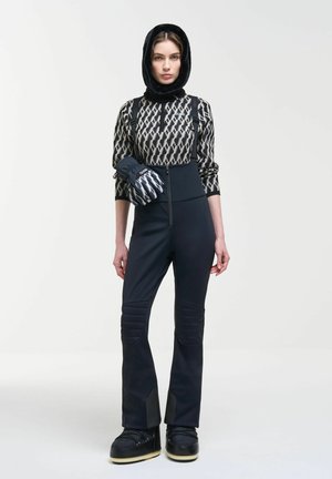 Femme portant un pantalon de ski noir matelassé avec bretelles, un haut à manches longues à motifs, une capuche noire et des gants d'hiver noirs tenant un poignet.