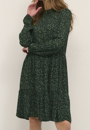 Frau in langärmeligem dunkelgrünen Blumenkleid, die mit einer Hand nahe am Gesicht vor einem einfarbigen hellen Hintergrund steht.