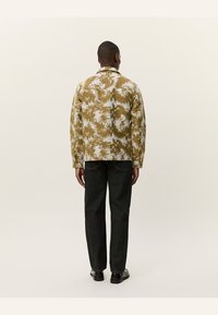Patterned jacket in brown and cream fabric with a relaxed fit. Pair of dark trousers and black shoes visible. Minimalist design overall.