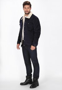 Námořnická kordurová bunda s krémovým sherpa límcem, obsahující dvě náprsní kapsy a zapínání na knoflíky, spárovaná s tmavými džínami a černými bojovými botami.