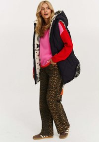 Frau in einem pinken und roten Pullover, schwarzer Kapuzenweste mit Futter aus Fell, Leopardenmuster-Hose und braunen Turnschuhen, die mit der Hand in der Tasche steht.