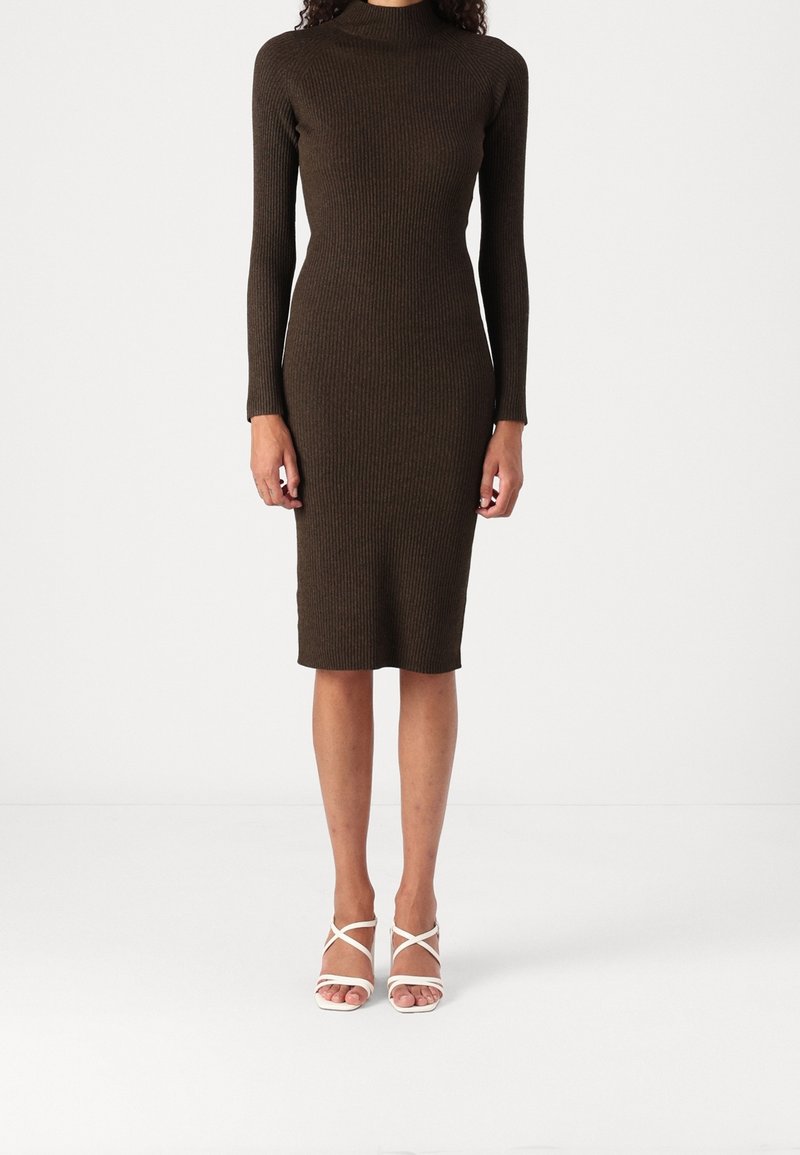 Femme portant une robe moulante marron foncé à manches longues et longueur genou avec des talons blancs à lanières et orteils ouverts sur fond blanc uni.