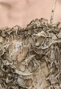 Κοντινό πλάνο ενός τοπ χωρίς μανίκια με βολάν, από ύφασμα με μπεζ και καφέ σχέδιο φίδι, στο άνω μέρος του στήθους και του ώμου ενός ατόμου.