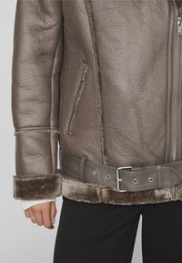 Giubbotto in pelle marrone con finitura testurizzata, caratterizzato da una vita con cintura e fodera in shearling ai polsini e alla parte inferiore, tasche con zip.