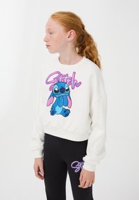 Felpa bianca corta con una grafica colorata di Stitch e testo. Abbinata a leggings neri con accenti grafici simili.