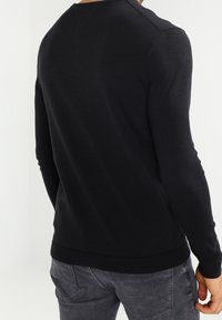 Pull noir à manches longues, coupe ajustée, fabriqué en matériau doux, avec un col rond et un ourlet côtelé, porté sur un pantalon gris foncé.