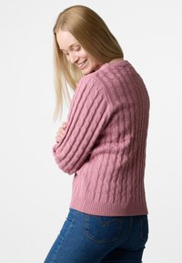 Kabelstrickpullover in sanftem Pink, mit strukturiertem Muster und geripptem Saum. Kombiniert mit blauen Jeans, die einen lässigen Sitz zeigen.