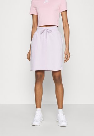 Mujer que lleva una camiseta corta rosa de Nike, falda con cordón color lila claro, calcetines blancos y zapatillas blancas, de pie frente a un fondo liso.