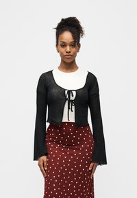 Γυναίκα με σγουρά μαλλιά δεμένα, φοράει μαύρο διαφανές μπλουζάκι με μακριά μανίκια και δέσιμο μπροστά πάνω από λευκό πουκάμισο και κόκκινη φούστα με λευκά μοτίβα καρδιών.