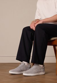 Grijze suède sneakers met een low-top ontwerp, witte zool en geprofileerde accenten, gecombineerd met zwarte broeken en witte sokken.