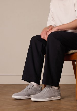 Grijze suède sneakers met een low-top ontwerp, witte zool en geprofileerde accenten, gecombineerd met zwarte broeken en witte sokken.