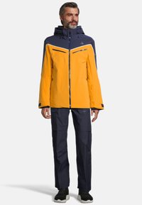 Wasserfeste gelbe und marineblaue Jacke mit einem Reißverschluss, verstellbarer Kapuze und mehreren Reißverschlusstaschen. Kombiniert mit marineblauen Hosen und schwarzen Schuhen.