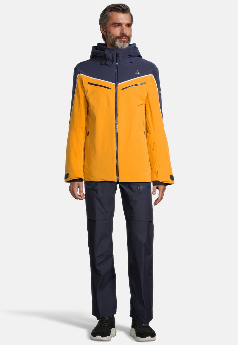 Wasserfeste gelbe und marineblaue Jacke mit einem Reißverschluss, verstellbarer Kapuze und mehreren Reißverschlusstaschen. Kombiniert mit marineblauen Hosen und schwarzen Schuhen.