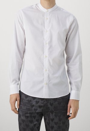 Ein Mann trägt ein weißes, langärmeliges Hemd mit Stehkragen und eine dunkle Hose mit Herzmuster.