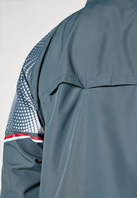 Graue Jacke mit glatter Textur, einem gemusterten Ärmeldesign in Blau und Weiß sowie einem roten Akzentstreifen.