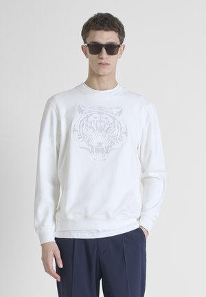 Sudadera blanca de algodón con un prominente gráfico de tigre gris en la parte delantera, puños y dobladillo acanalados, combinada con pantalones azul marino.