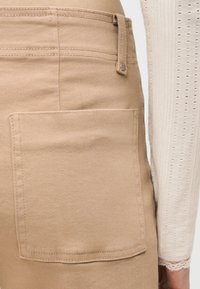 Μπεζ φούστα με ορατή πίσω τσέπη και θηλιά για ζώνη, φορεμένη με κρεμ μακρυμάνικη μπλούζα που έχει μικρό διάτρητο μοτίβο και δαντελένιο τελείωμα στην μανσέτα.