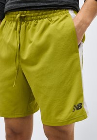 Άνδρας που φοράει αθλητικά σορτς σε λαχανί χρώμα με λευκές πλαϊνές λωρίδες και μαύρο λογότυπο New Balance, με το χέρι στην τσέπη, με γκρι πουκάμισο που φαίνεται μερικώς.