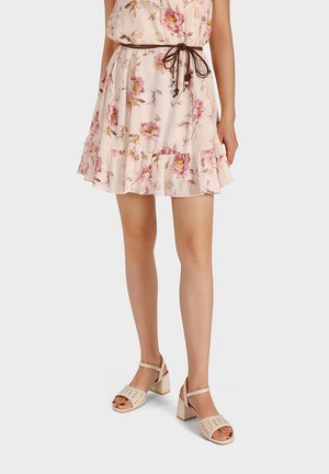 Donna che indossa un vestito floreale rosa chiaro con una cintura marrone annodata e sandali beige con tacco a blocco e punta aperta, su uno sfondo semplice.