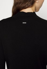 Czarny prążkowany sweter z wysokim kołnierzem i charakterystycznym srebrnym logo "BOSS" na plecach. Materiał ma miękką fakturę.