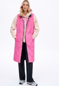 Różowa kamizelka pikowana w romby, z granatowym kołnierzem, przednimi kieszeniami i długim krojem, noszona na beżowej bluzie z kapturem.