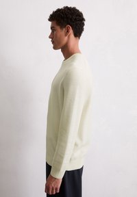 Hellgelb strukturierter Pullover mit Rundhalsausschnitt und langen Ärmeln, über dunklen Hosen getragen, von der Seite vor einem neutralen Hintergrund gezeigt.