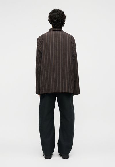 Άτομο με σγουρά μαλλιά, γυρισμένο πλάτη, φοράει σκούρο καφέ σακάκι με ρίγες και φαρδιά μαύρη παντελόνια σε μονόχρωμο φόντο.