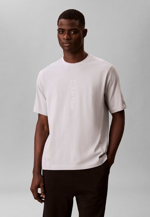 Mand iført en lysegrå Calvin Klein t-shirt med korte ærmer og sorte bukser, stående foran en ensfarvet grå baggrund.