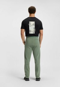 Schwarzes T-Shirt mit kurzen Ärmeln und einem grafischen Druck auf der Rückseite, kombiniert mit grünen Sporthosen. Die Hosen haben eine glatte Textur und eine taillierte Passform.
