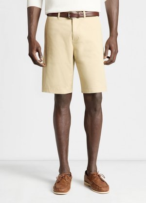 Uomo che indossa pantaloncini beige a lunghezza ginocchio con cintura, scarpe in pelle marrone con lacci e una maglia bianca a maniche lunghe su sfondo bianco.