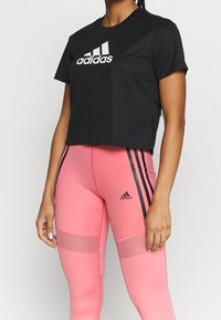 Černé krátké tričko s bílým logem „adidas“, doplněné růžovými legínami s černými pruhy a síťovanými panely.