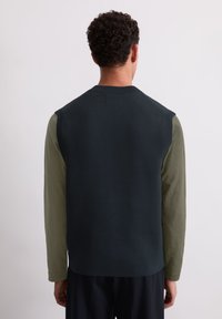 Dunkelblauer, ärmelloser Pullover über einem langärmeligen olivgrünen Hemd, mit einem gerippten Halsausschnitt und klaren Linien, ohne sichtbare Muster.