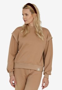 Beżowy sweatshirt z opadłymi ramionami, prążkowanymi mankietami i teksturowaną naszywką z logo na dole, wykonany z miękkiego, gładkiego materiału.